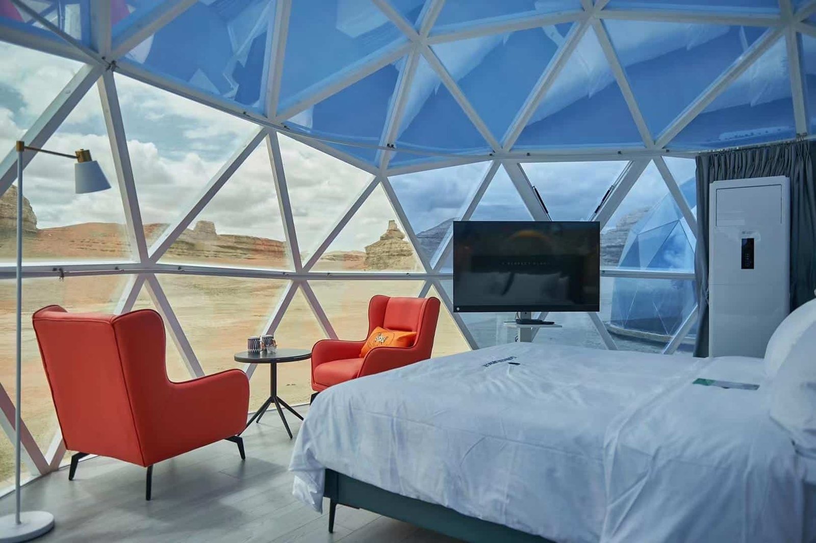 Modern dome shaped room with large triangular windows, two red chairs, a round table, a floor lamp, a tv, and a bed, overlooking a desert landscape with rock formations.