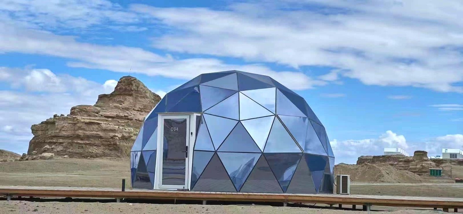 A geometric dome shaped structure with reflective panels sits on a wooden platform in a desert landscape with rock formations and a partly cloudy sky.