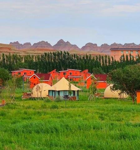 A green field with tents and small red houses, trees in the background, and rocky hills under a cloudy sky.