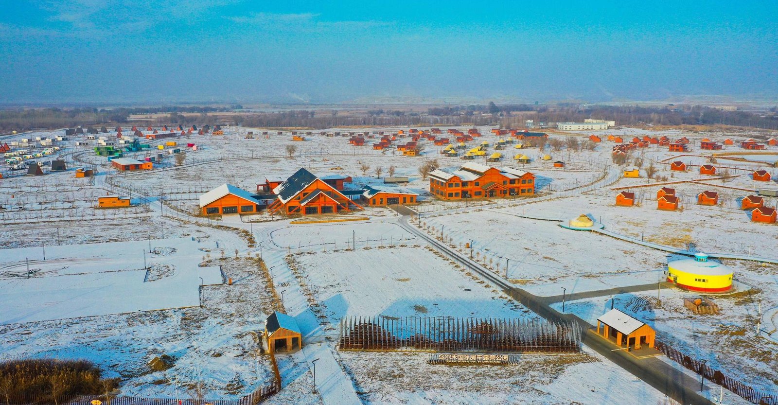 Aerial view of a snowy landscape with scattered orange buildings, small houses, and clear roads under a blue sky.