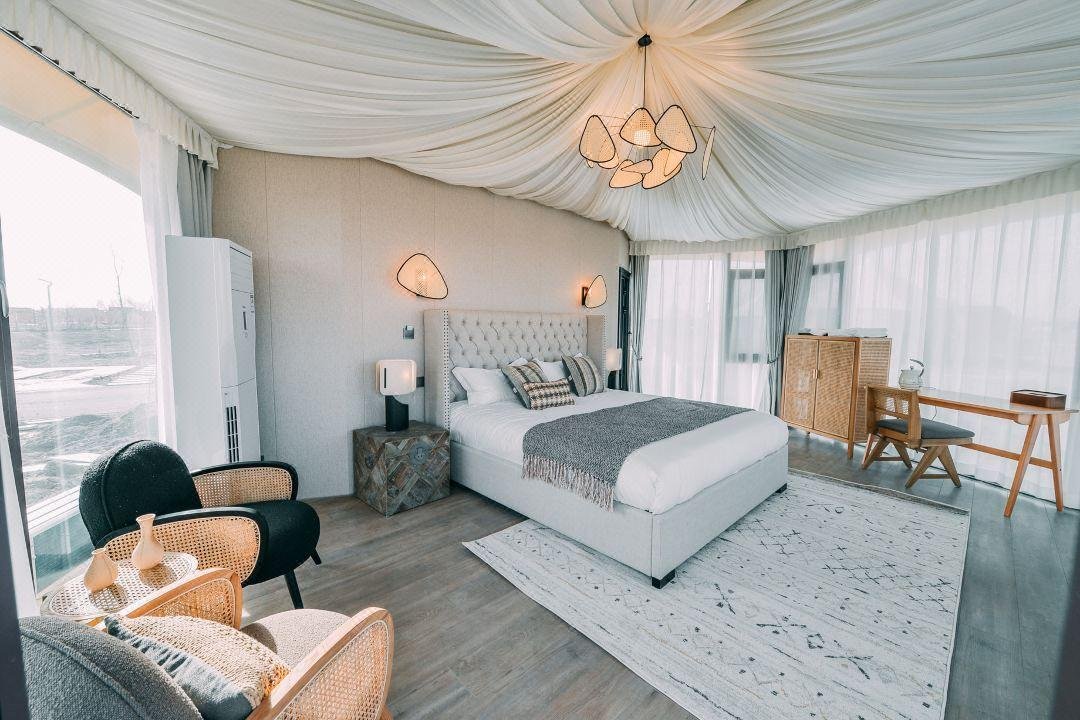 Modern bedroom with a canopy ceiling, upholstered bed, armchairs, desk, and large windows with sheer curtains, featuring neutral tones and natural light.