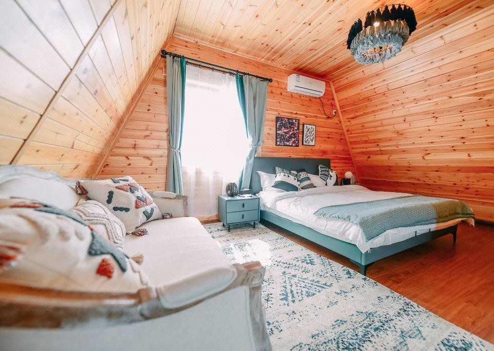 A cozy attic bedroom with wood paneled walls, a bed, a sofa, blue accents, a rug, and a window with turquoise curtains letting in natural light.