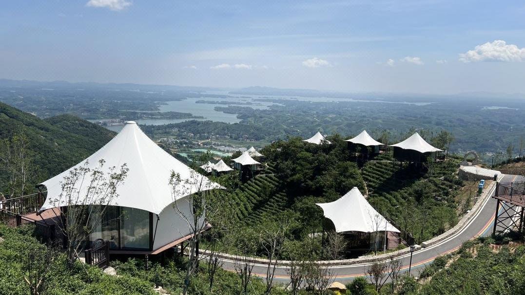 White tent like structures are arranged on terraced hillsides overlooking a winding road, lush greenery, and a distant view of a lake and mountains under a clear sky.