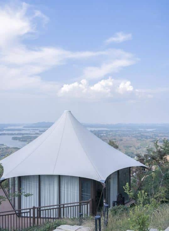 A white, tent like structure with large windows stands on a hillside overlooking a scenic landscape with water and distant mountains under a partly cloudy sky.