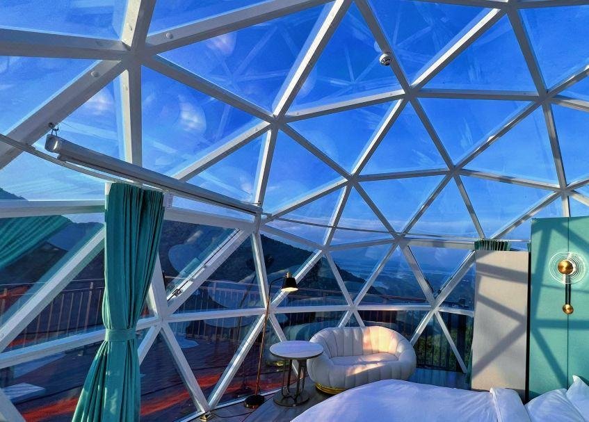 A geodesic dome room with glass panels, modern furniture, blue curtains, and mountain views visible through the transparent walls and ceiling.