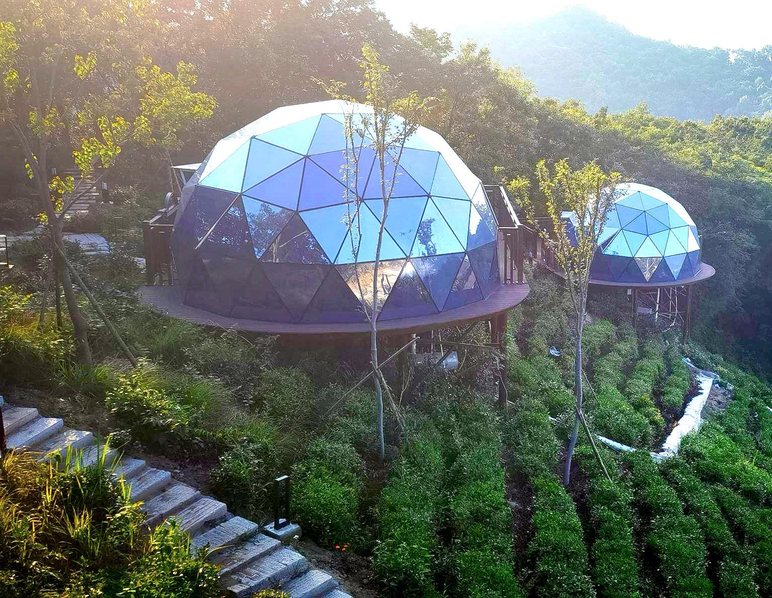 Two glass geodesic dome structures are elevated on platforms in a lush, terraced hillside surrounded by greenery and trees.
