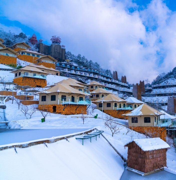 Snow covered hillside resort with multiple wooden cabins, terraced along the slope, surrounded by snowy trees and under a partly cloudy blue sky.