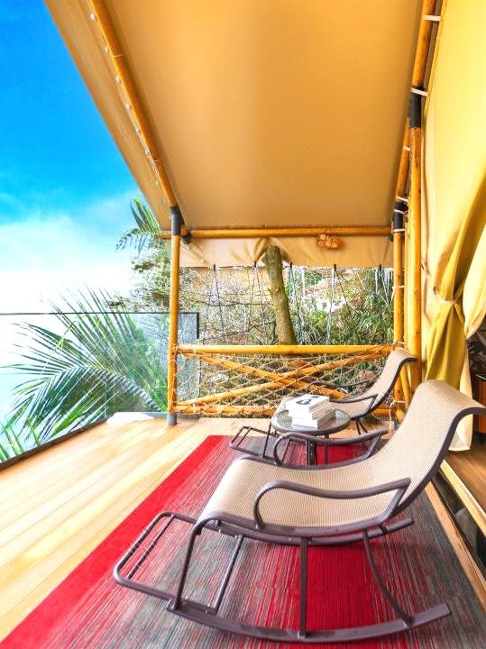 Two mesh lounge chairs and a small table sit on a covered wooden balcony with a red rug, overlooking trees and a blue sky.