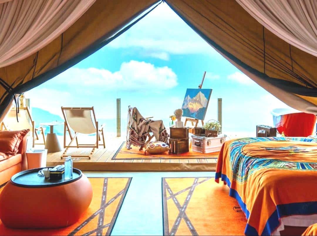 A glamping tent interior with bright bedding, rugs, and a seating area, opening to a deck with chairs, an easel, and scenic mountain views under a blue sky.