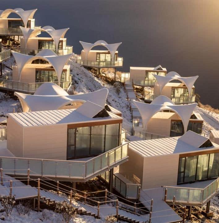 Modern white buildings with curved roofs and glass balconies are arranged on a snowy hillside at sunset.