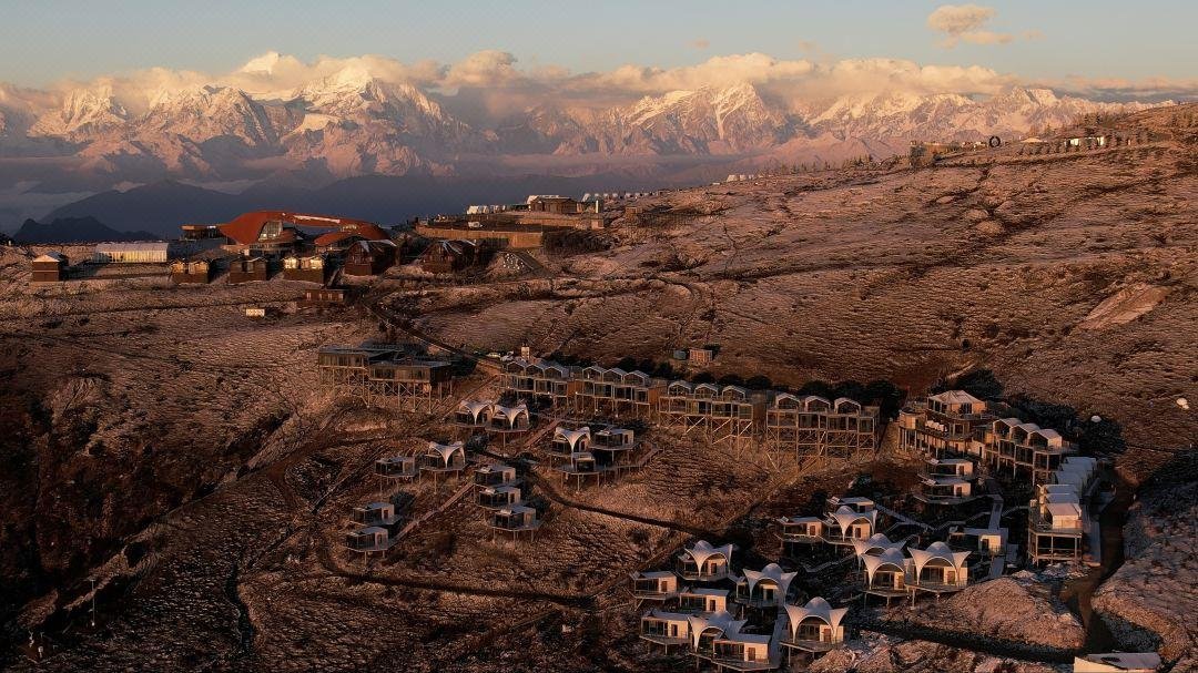 A mountainside resort with clustered cabins and lodges, set against a dramatic backdrop of snow capped peaks under a partly cloudy sky at sunset.