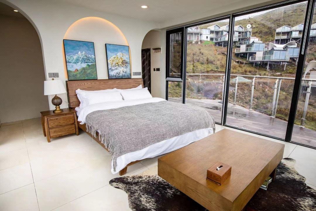 Modern bedroom with a wooden bed, gray blanket, bedside table, two mountain paintings, large windows, and a view of hillside houses.