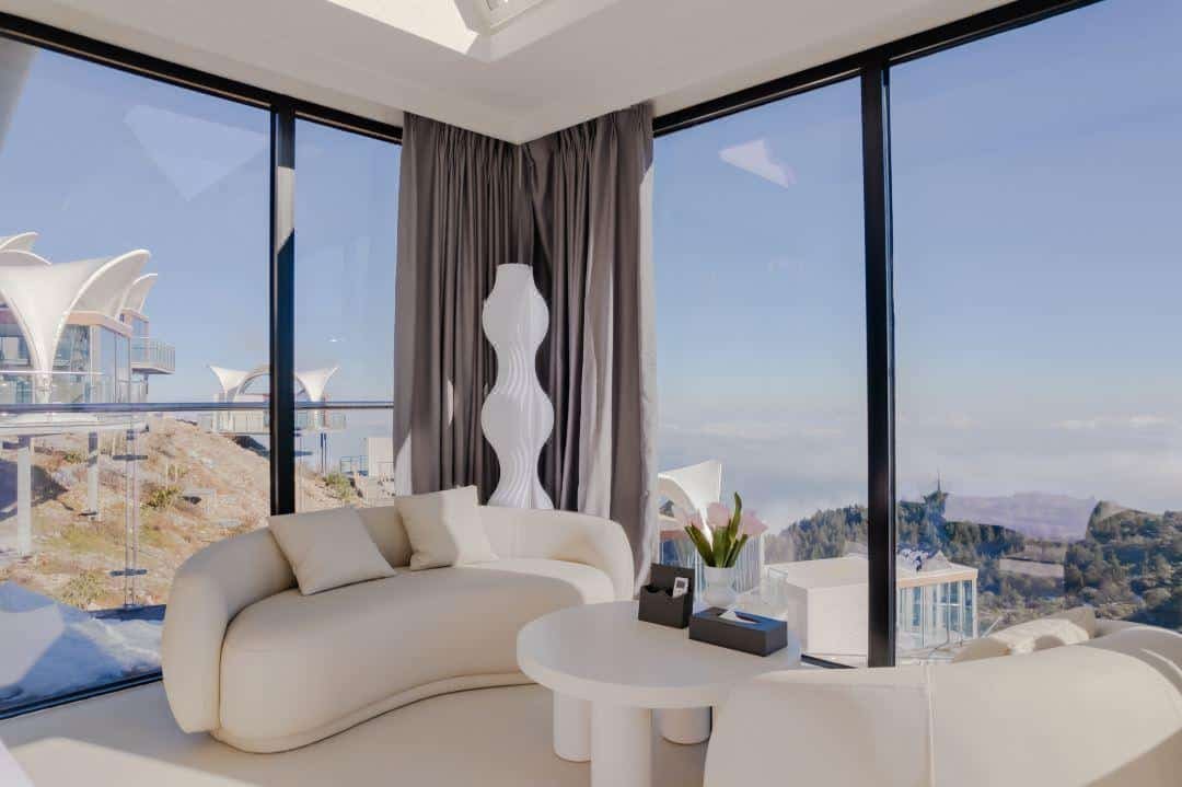 Modern living room with white curved sofa, floor lamp, and large windows offering a scenic mountain view under a clear sky.