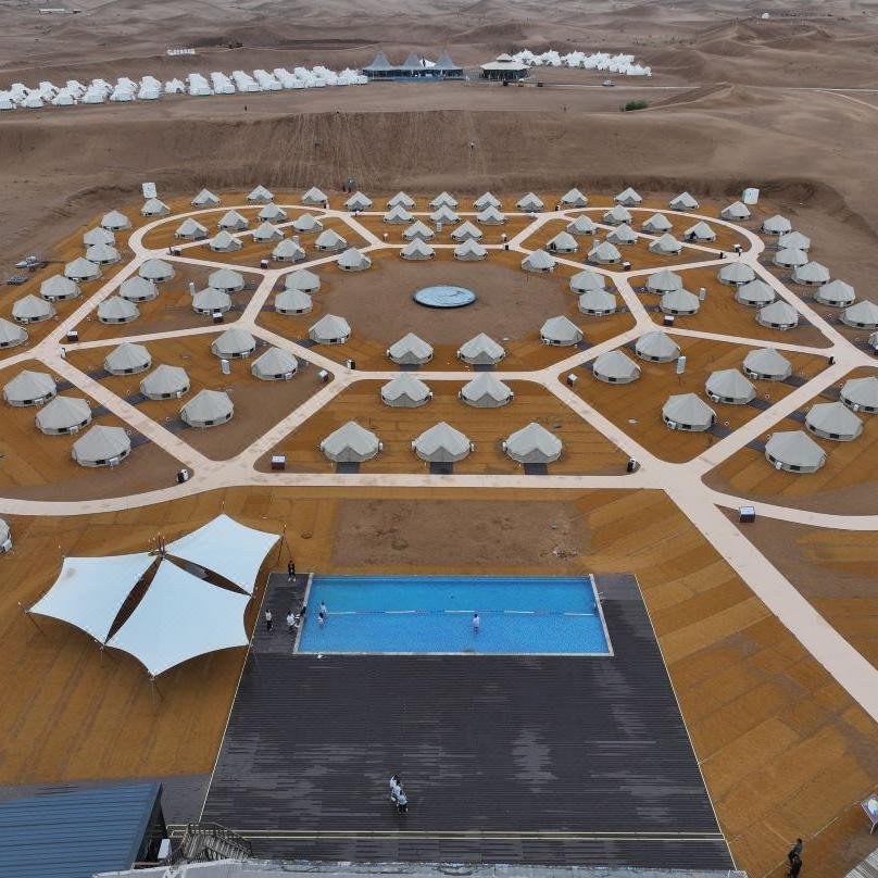 Aerial view of a desert glamping site with circular arrangement of tents, a central fountain, and a swimming pool in the foreground.