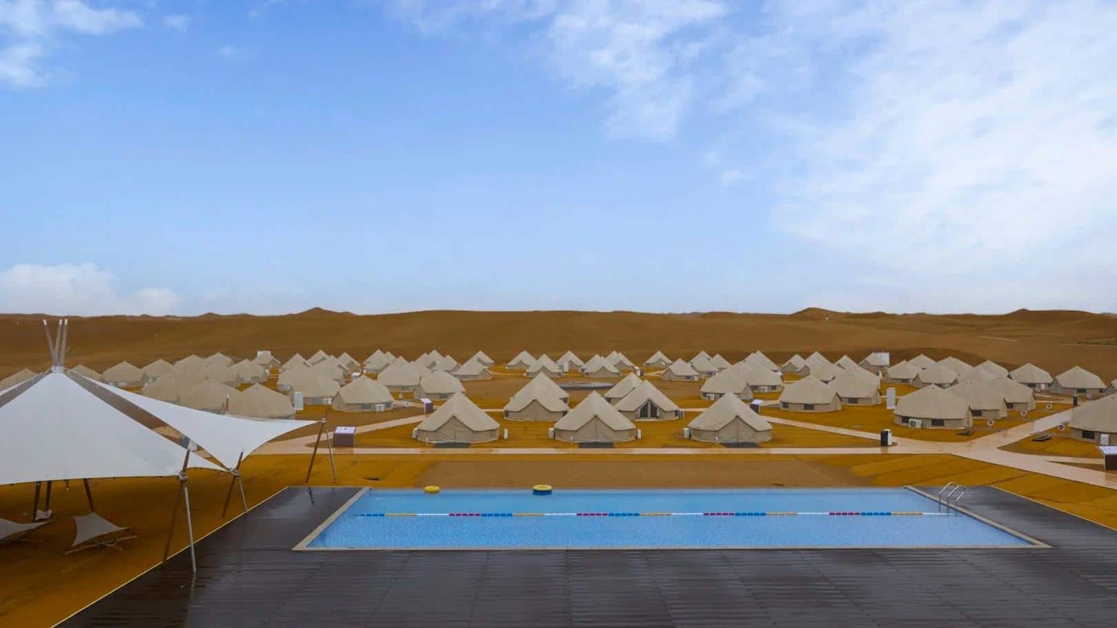 Rows of beige tents are set up on sandy terrain behind a rectangular outdoor swimming pool under a blue sky with scattered clouds.