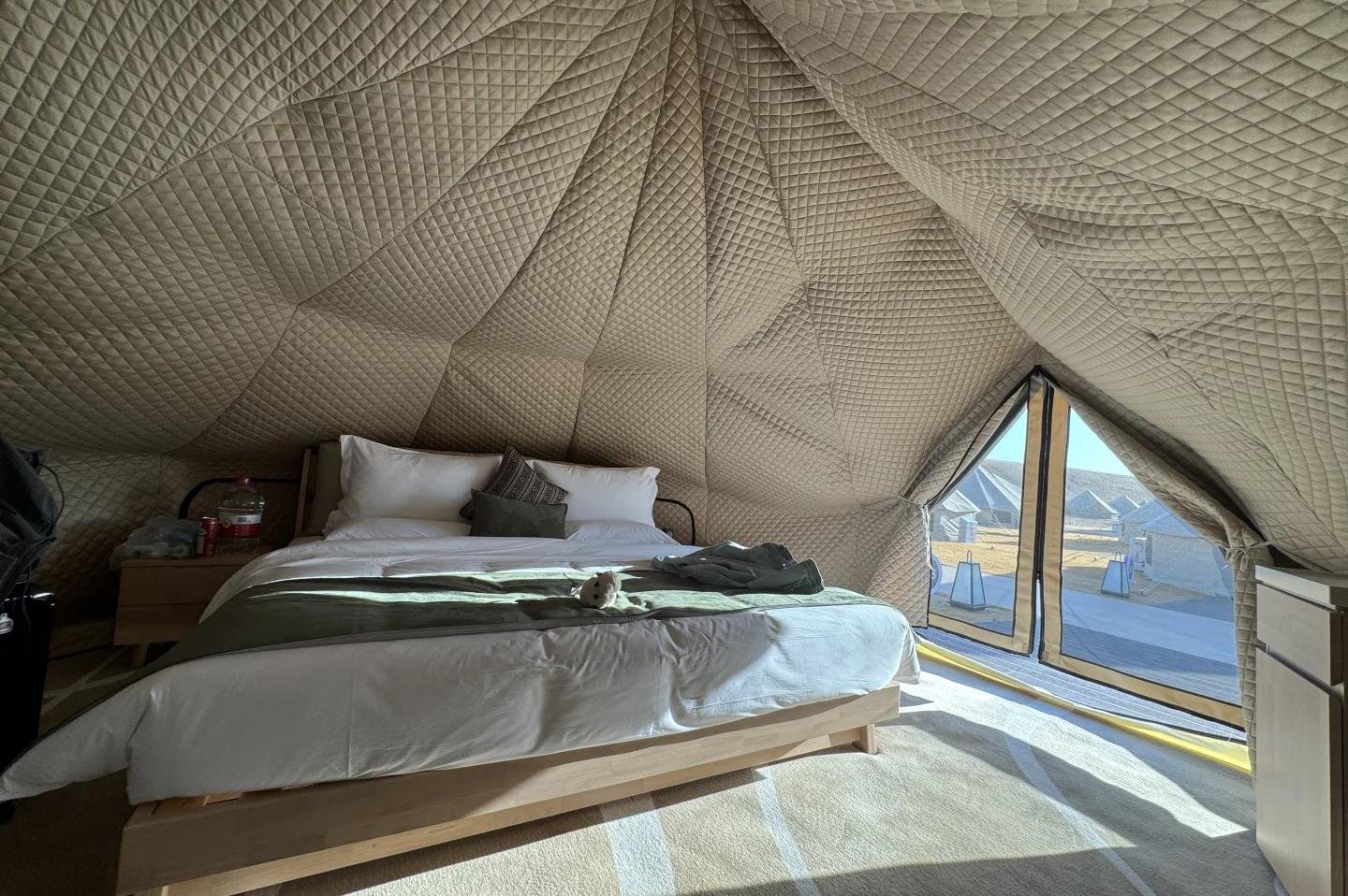 Bed inside a dome shaped tent with quilted walls, large windows letting in sunlight, and simple wooden furniture.