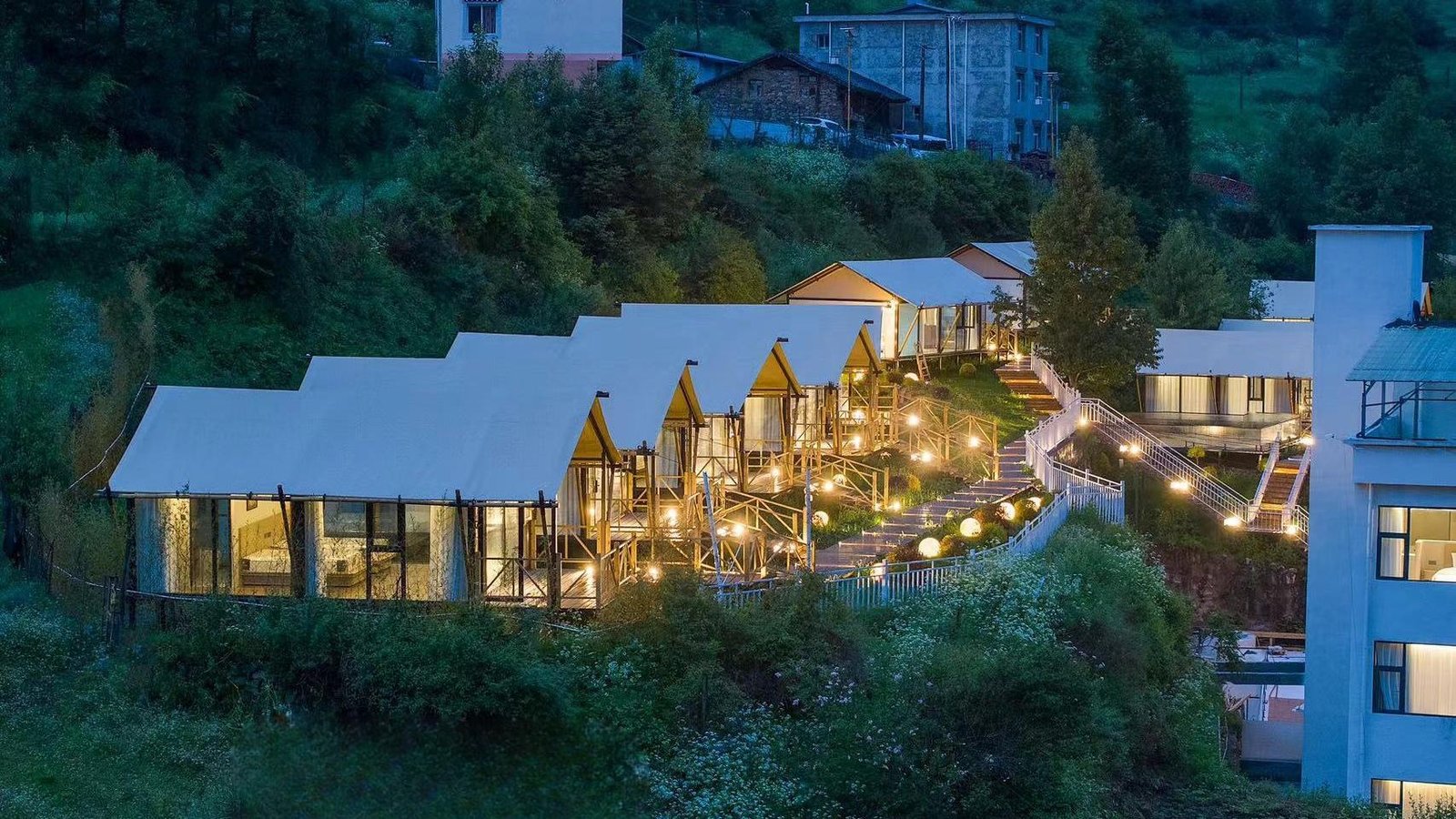 A row of modern cottages with large windows and outdoor lights is set on a hillside surrounded by greenery and other buildings in the background.