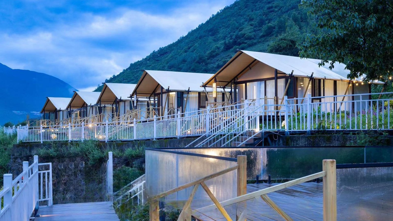 Row of modern glamping tents with white railings set on a hillside, surrounded by greenery and mountains under a cloudy sky.